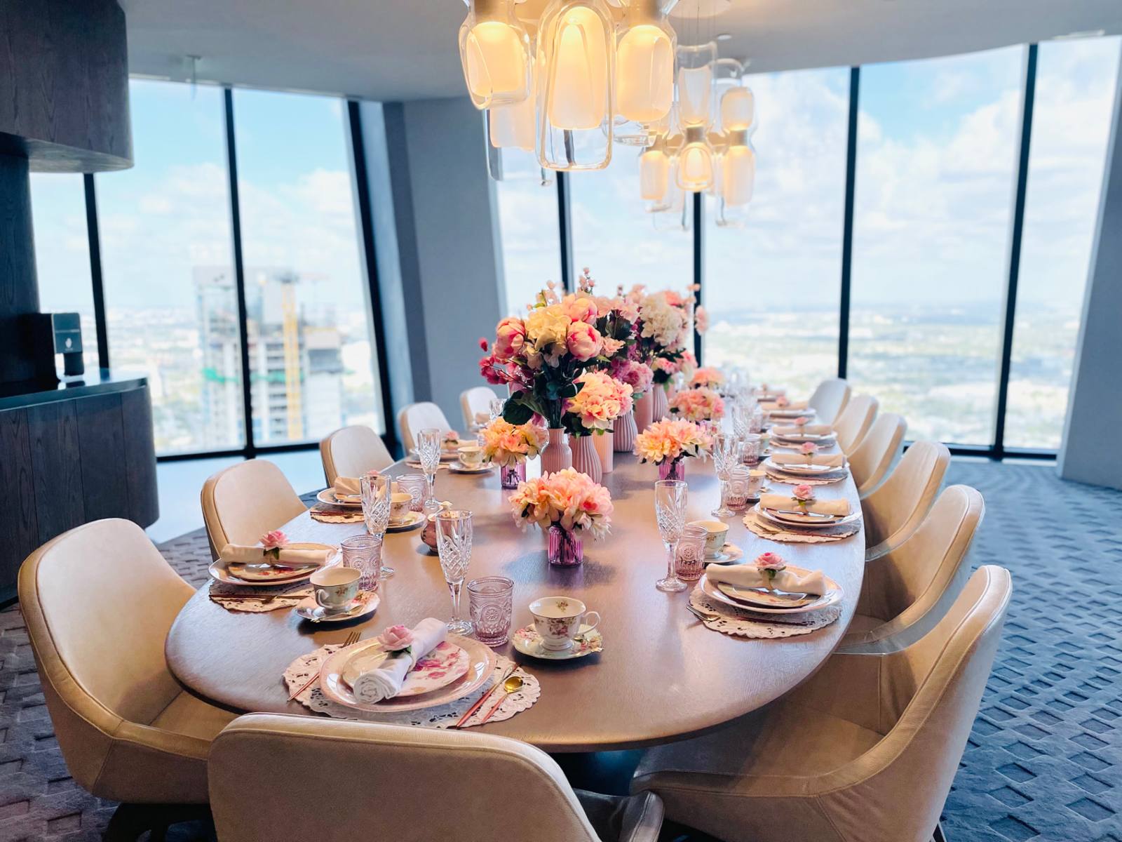 A long oval tea table set with pink florals and floral china in a Miami high-rise dining room with panoramic city views